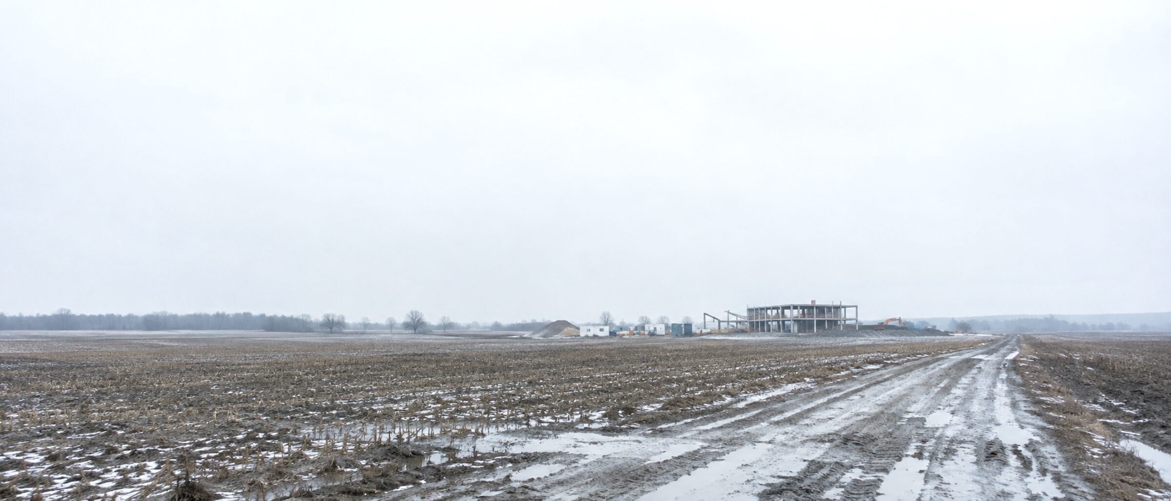Weite Feldflur, ein Feldweg führt zu einem einzelnen Rohbau am Horizont